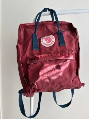 Fjällräven Kånken Backpack - Burgundy/Blue - Classic Fjällräven Kånken backpack in a beautiful burgundy colour with blue shoulder straps. In good pre-loved condition with no major flaws. Clean, durable and perfect for school, work, travel or everyday use. A timeless Scandinavian design that never goes out of style. Spacious main compartment, front zip pocket and comfortable carrying handles. ✔️ Good condition  ✔️ Original Fjällräven Kånken  ✔️ Trendy colour combination  ✔️ Ready to use