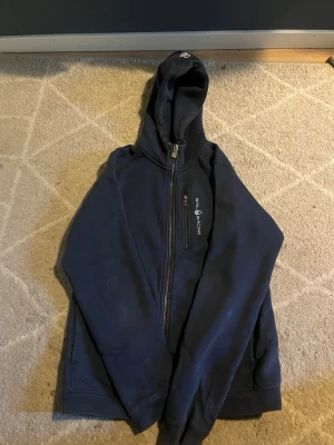 Mörkblå zip-hoodie med huva - Mörkblå hoodie med dragkedja och huva. Tryckt logga på bröstet och två fickor framtill. Perfekt för en avslappnad stil och enkel att matcha med jeans eller joggers.