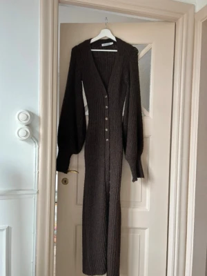 Brun Adoore ribbad långklänning med knappar - Mysig brun långklänning med ribbad struktur och långa puffärmar. Klänningen har v-ringning och knappar framtill, vilket ger en snygg och stilren look. Perfekt för dig som gillar en figurnära passform och vill ha något både bekvämt och trendigt.