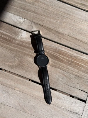 Svart klocka från Daniel Wellington - Stilren svart klocka från Daniel Wellington med rund urtavla och svart läderarmband. Modell Classic Sheffield Silver. Sparsamt använd, inga skador.  Nypris: 1 999kr Säljes för: 450kr