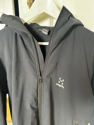 Svart vindjacka från Haglöfs - Snygg svart vindjacka från Haglöfs med huva och hel dragkedja framtill. Jackan har minimalistisk design, två sidofickor med dragkedja och diskret logga på bröstet. Tillverkad i ett lätt och slitstarkt material som passar perfekt för blåsiga dagar. 