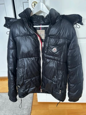 Moncler Black Puffer Jacket - Det här är en svart Moncler-pufferjacka med huva. Jackan har en glansig yta, dragkedja framtill, sidofickor med dragkedja samt en bröstficka med Moncler-logotypen. Jackan har dragskor både i nederkanten och i huvan för justering. Baksidan är enkel med horisontell quiltning. Skicket verkar vara varsamt använt och det finns inga synliga större defekter. 