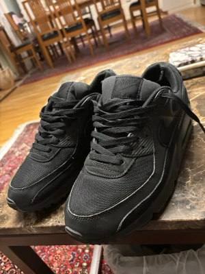 Svarta Nike Air Max sneakers - Säljer ett par helsvarta Nike Air Max sneakers med klassisk siluett. Skorna har mesh och syntetmaterial i ovandelen, samt en chunky sula med synlig Air-enhet. Swoosh-logga på sidan och vadderad plös för extra komfort.