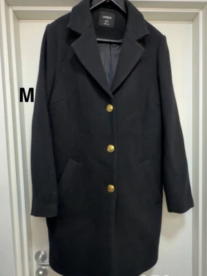 Svart kappa med guldknappar Lindex - Stilren svart kappa från Lindex med tre guldknappar framtill och klassisk krage. Kappan har två snedställda fickor. Perfekt för dig som vill ha en enkel men snygg look till hösten. 🌸 Sparsamt använd. Ej tvättad. Storlek m