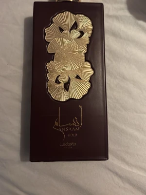 Ansam Gold parfym från Lattafa - Lyxig parfymflaska i mörkbrunt med guldfärgade blommönster och elegant design. Flaskan har en rektangulär form och detaljer i guld som ger en exklusiv känsla. Perfekt för dig som gillar unika och snygga parfymflaskor.