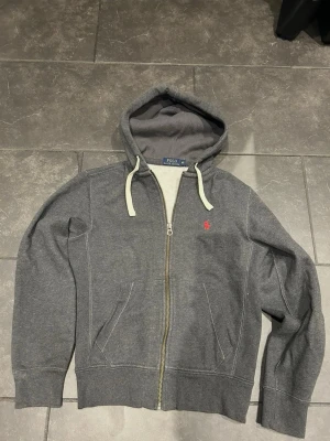Grå hoodie från Polo Ralph Lauren - Mörkgrå hoodie med dragkedja från Polo Ralph Lauren. Klassisk design med vit dragsko i huvan och röd broderad logga på bröstet. Tillverkad i mjukt bomullsmaterial och har två fickor framtill. Perfekt för en avslappnad och stilren look.
