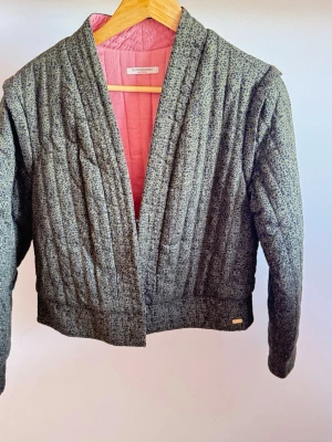 Mönstrad quiltad cardigan från Scotch & Soda Strl M - Quiltad cardiganjacka från Scotch & Soda med grått och svart småmönstrat tyg och rosa innerfoder. Jackan har öppen front utan knappar, lång ärm och bred fåll. Perfekt för dig som gillar unika detaljer och skön passform.