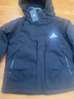 Svart vindjacka med huva från Adidas - Snygg svart vindjacka från Adidas med huva och dragkedja framtill. Jackan har två stora fickor med dragkedja och Adidas-logga på bröstet. Perfekt för blåsiga dagar och sportiga outfits.