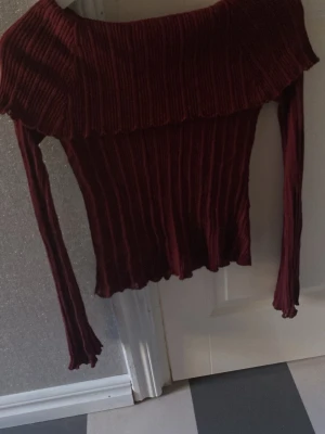 Vinröd off shoulder tröja - Trendig vinröd ribbad tröja med off shoulder-design och långa ärmar. Tröjan har en bred volangkant upptill och är tillverkad i ett stretchigt material som sitter snyggt på kroppen. Perfekt för dig som gillar en stilren men ändå unik look.