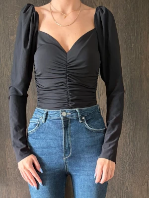 Svart body med puffärmar - Trendig svart body med långa puffärmar och djup v-ringning. Framsidan har snygg rynkning som framhäver midjan. Tillverkad i stretchigt material som sitter bekvämt och smickrande.