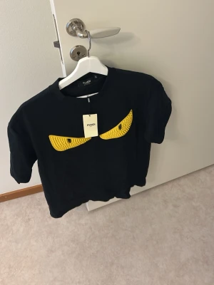 Svart Fendi t-shirt med gula ögon - Cool svart t-shirt från Fendi med ikoniska gula ögon framtill. T-shirten har rund hals och korta ärmar. Perfekt för dig som vill sticka ut med en unik och trendig design.