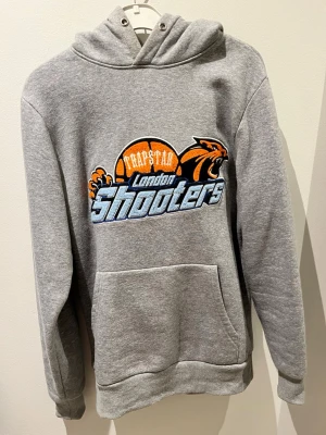 Grå hoodie Trapstar London Shooters - Snygg grå hoodie med stor broderad Trapstar London Shooters-logga på bröstet. Hoodien har huva, känguruficka och ribbade muddar. Trycket har orange, svart och blå detaljer med baskettema och pantermotiv. Perfekt för en sportig streetwear-look.  Den är använd 4gånger, har du mer frågor så tveka inte att höra av dig👍🏽