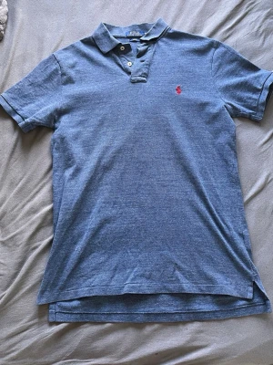 Polo Raulph Lauren piké - Klassisk blå Polo Raulph Lauren piké med krage och knappar framtill. Tröjan har en liten röd broderad logga på bröstet och är kortärmad. Perfekt till jeans eller shorts för en avslappnad look.