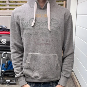 Grå SuperDry Hoodie - Säljer en grå hoodie från Superdry med tryck på framsidan. Den har en klassisk känguruficka och justerbar huva med snören. Perfekt för en avslappnad stil.