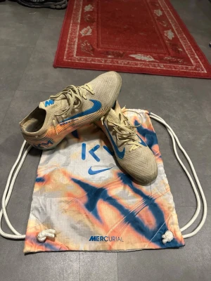 Nike Mercurial beige/blå fotbollsskor - Säljer ett par Nike Mercurial fotbollsskor i orange med blå detaljer och orange inslag. Skorna är i FG donbar. De är storlek 44. Skorna har en strömlinjeformad design, låg profil och mönstrad sula för bra grepp på planen. Medföljer en matchande Mercurial-påse i vitt, blått och orange.