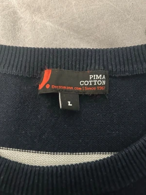 Marinblå randig stickad tröja L - Snygg marinblå stickad tröja från Dressmann i storlek L, tillverkad i mjuk Pima bomull. Tröjan har vita horisontella ränder och rund halsringning med ribbade muddar. Perfekt för en stilren och avslappnad look.