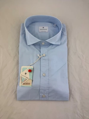 Ljusblå skjorta från Bastoncino - Säljer en klassisk ljusblå skjorta från Bastoncino, tillverkad i Italien. Skjortan har en stilren design med knappar framtill och normal passform. Perfekt till både jeans och kostymbyxor. Materialet känns mjukt och svalt.