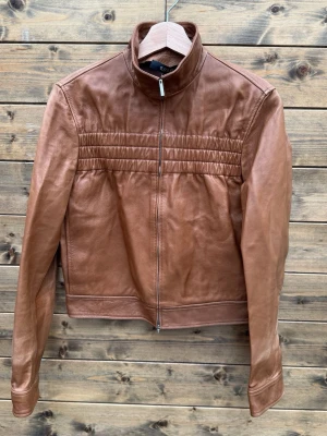 Gucci Brown Leather Jacket (M-L) - These images show a brown leather jacket displayed on a wooden hanger against a wooden background. The first image is the front view, featuring a zip-up closure, a stand collar, and horizontal stitched detailing across the chest. The second image is the back view, showing clean lines and simple stitching, with a slightly curved hem at the back. The jacket has a classic, minimalist design with long sleeves and zippered cuffs.