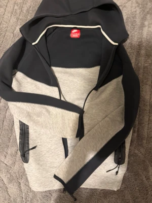 Nike tech  - Snygg hoodie i grått och svart med dragkedja framtill och huva. Jackan har svarta detaljer på ärmar och axlar samt praktiska fickor med dragkedja. Perfekt för träning eller chill. Materialet ser ut att vara mjukt och bekvämt. Har inte använt den så mycket. 