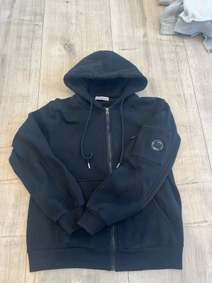 Svart hoodie med dragkedja och linsdetalj - Säljer en svart hoodie från C.P. Company med dragkedja framtill och klassisk huva med dragsko. På vänster ärm finns den ikoniska linsen och en ficka. Hoodien har ribbade muddar och är tillverkad i mjukt bomullsmaterial. Perfekt för en avslappnad streetwear-look.