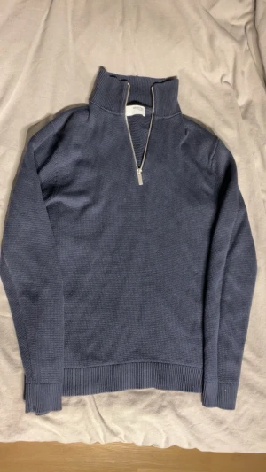 Blå stickad halvzip tröja - Mörkblå stickad tröja med halvzip och hög krage. Tröjan har långa ärmar, ribbade muddar och nederkant. Perfekt för lager på lager och chill stil. Materialet ser ut att vara bomull eller bomullsblandning.