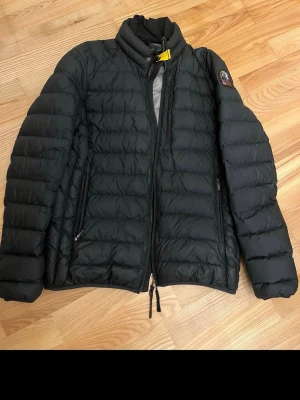 Svart dunjacka från Parajumpers - Snygg svart dunjacka från Parajumpers med quiltad design och hög krage. Jackan har två dragkedjeförsedda fickor framtill och detaljer i gult vid halsen. Insidan är grå med orange detaljer och logotyp på ärmen.