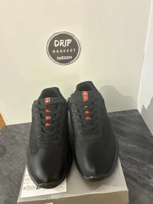 Svarta Prada sneakers med röd detaljer.  - Snygga svarta sneakers från Prada i storlek 45, med diskret röd logga på plösen. Skorna har en låg silhuett, svarta skosnören och en mix av läder och meshmaterial för en modern look. Perfekta för dig som vill ha en clean och exklusiv stil.