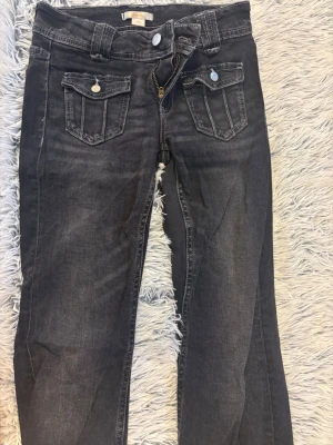 Svarta jeans med fickdetaljer - Snygga svarta jeans med slitna detaljer och två framfickor med lock och knapp. Jeansen har raka ben, bälteshällor och klassiska bakfickor. Materialet är denim med en tvättad look som ger en cool vibe.
