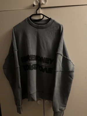 Oversized grå sweatshirt med tryck - Cool oversized grå sweatshirt med svart texttryck på bröstet. Tröjan har synliga sömmar och nedhasade axlar för en avslappnad streetwear-look. Mjuk bomullskänsla och ribbad mudd vid hals, ärmslut och nederkant.