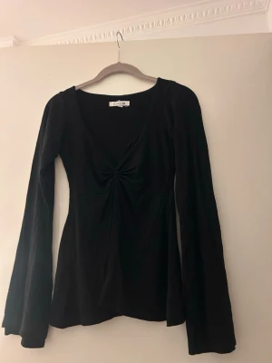 Svart långärmad topp med twistdetalj - Snygg svart långärmad topp med v-ringning och twistdetalj framtill. Ärmarna är vida och materialet känns mjukt och stretchigt, perfekt för en trendig look. Passformen är normal och toppen är enkel att styla till olika outfits.