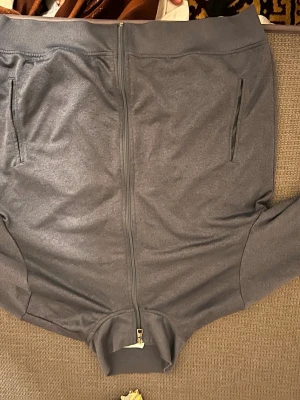 Mörkgrå ziptröja från EA7 - Snygg mörkgrå ziptröja från EA7 Emporio Armani med hel dragkedja framtill och två sidofickor med dragkedja. Tröjan har en enkel och stilren design med ribbade muddar vid ärmslut och nederkant. Perfekt för en avslappnad och sportig look.