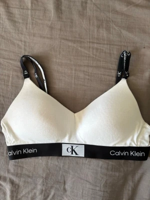 Vit Calvin Klein bralette - Vit bralette från Calvin Klein med svarta, justerbara axelband och bred resår med logga. Mjuk och utan bygel, perfekt för en avslappnad stil. Axelbanden har tryckt CK-logga och resåren ger bra stöd. Materialet känns lent och stretchigt.