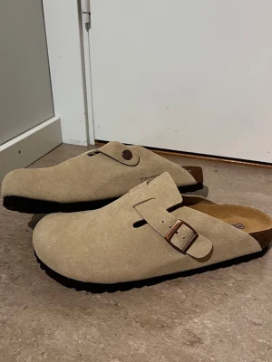 Beige mockatofflor från Birkenstock - Säljer ett par snygga beige tofflor från Birkenstock i mocka med justerbart spänne i metall. De har en klassisk slip-in design och mjuk innersula för extra komfort. Perfekta för chill dagar hemma eller när du vill ha något bekvämt på fötterna.
