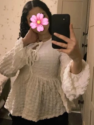 Vit volangblus med knytning - Supersöt vit blus med volangiga, vida ärmar och knytning fram i halsen. Blusen har en strukturerad, krinklad yta och en lös, flowy passform som ger en romantisk vibe. Perfekt att styla med jeans eller kjol för en trendig look.