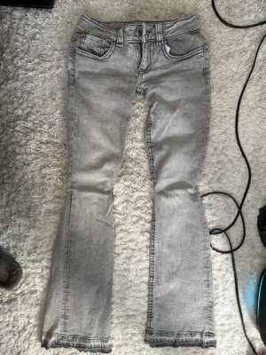 Grå bootcut jeans med fickdetaljer - Säljer ett par ljusgrå bootcut jeans med slitna detaljer och snygga fickor med lock och knapp bak. Jeansen har klassisk femficksdesign, bälteshällor och dragkedja med knapp framtill. Perfekta för en avslappnad och trendig look. Low waist för dig som gillar en y2k look.