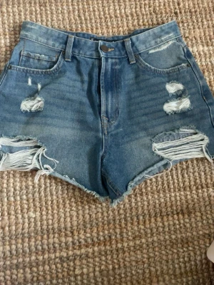 Högmidjade blå jeansshorts - Snygga högmidjade blå jeansshorts med slitna detaljer och råa kanter. Klassisk femficksmodell med bälteshällor och dragkedja fram. Perfekta för sommaren och ger en avslappnad vibe.