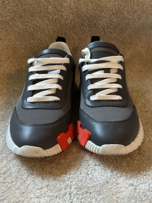 Grå sneakers med vita och orange detaljer - Snygga sneakers i grått med vita snören och sula. Skorna har coola detaljer i orange och svart på sulan samt dragflik bak. Ovandelen är i mesh och syntetmaterial för en modern look. Perfekta för dig som gillar stilrena men ändå unika sneakers.