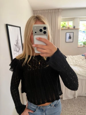 Svart spetsblus med volangdetaljer - Säljer en svart blus i spets med volangdetaljer på axlarna och långärmad design. Blusen har en lätt transparent look och är croppad, vilket ger en trendig vibe. Perfekt att styla med jeans eller kjol för en cool outfit.
