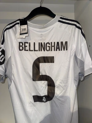Real Madrid Bellingham matchtröja - Säljer en vit Real Madrid matchtröja från Adidas med Bellingham och nummer 5 på ryggen. Tröjan har svarta detaljer, klubbmärke på bröstet, gula linjer längs sidorna och Adidas-logga. Tillverkad i funktionsmaterial, perfekt för fotboll eller träning.