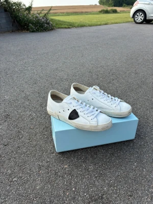 Feta Philippe modell skor i fint skick - Snygga vita sneakers i lågt snitt med svart detalj på sidan. Skorna har klassisk snörning och är tillverkade i läder med vit sula. Perfekta för dig som gillar en clean och stilren look med en twist.