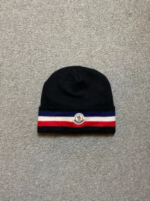 Svart Moncler mössa med ränder - Snygg svart mössa från Moncler med breda blå, vita och röda ränder längst ner samt Moncler-logga framtill. Klassisk modell i mjukt stickat material som håller dig varm och stilren under kalla dagar.