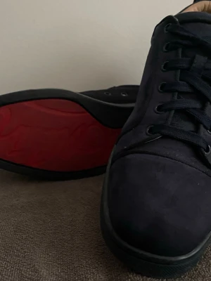 Mörkblå Louboutin’s med röd sula - Snygga mörkblå Loubs i mockaliknande material med svarta skosnören och svart sula. Insidan är beige och yttersulan har en cool röd detalj med mönster. Klassisk låg modell som passar till det mesta.