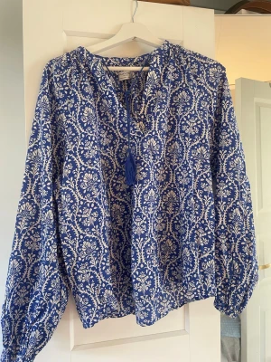 Blåvit blus från Holly & Whyte - Snygg blåvit blus från Holly & Whyte by Lindex med ett detaljerat blommönster. Blusen har långa ärmar med resår vid handlederna och knytband med tofsar vid halsen. Tillverkad i ett lätt och luftigt material, perfekt för en trendig look.