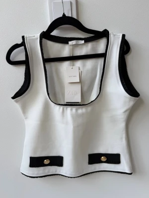 Topp från Zara - Stilren vit topp från Zara med svarta kontrastkanter och dekorativa svarta detaljer med guldfärgade knappar framtill. 