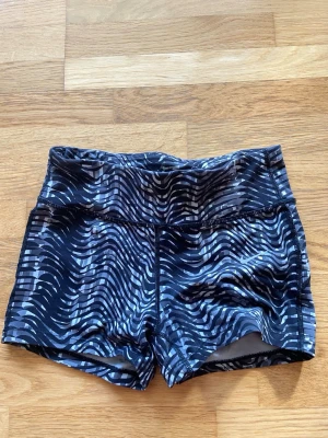 Nike Dri-FIT svarta träningsshorts - Säljer ett par svarta Nike Dri-FIT träningsshorts med hög midja och ett vågigt, randigt mönster i vitt och grått. Shortsen är tillverkade i ett stretchigt syntetmaterial som andas och passar perfekt för intensiva träningspass. Använd ganska mycket men är i bra skick. 