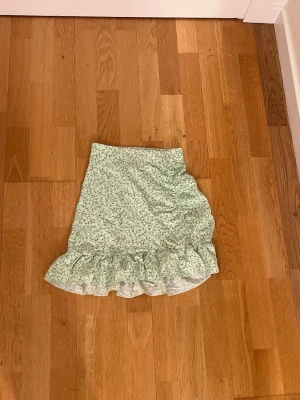 Ljusgrön blommig kjol med volang - Söt ljusgrön kortkjol med småblommigt mönster och volang längst ner. Kjolen har en feminin och somrig look, perfekt för dig som gillar en romantisk stil. Tillverkad i ett lätt och luftigt material som känns skönt mot huden.