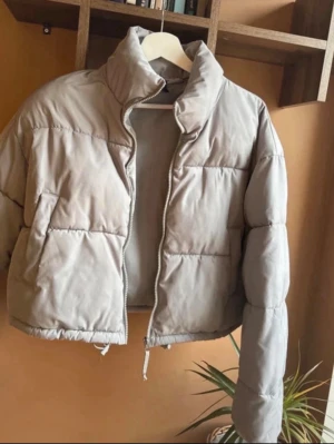 Beige cropped pufferjacka - Säljer en beige cropped pufferjacka med hög krage och dragkedja framtill. Jackan har en boxig passform och breda kanaler som ger en riktigt trendig look. Perfekt för kalla dagar och enkel att styla till olika outfits.