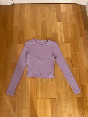 Ljuslila långärmad topp - En stilren ljuslila långärmad topp med croppad passform. Toppen har rund halsringning och är tillverkad i ett mjukt, stretchigt material som sitter snyggt på kroppen. Perfekt för att skapa en trendig och enkel look.