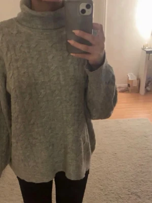Grå stickad polotröja - Mysig grå kabelstickad polotröja med oversized passform och långa ärmar. Tröjan har en hög polokrage och är perfekt för kalla dagar. Stickningen ger en klassisk och trendig look.
