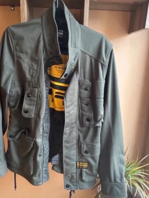 Olivgrön jacka från G-Star RAW - Snygg olivgrön jacka från G-Star RAW med flera fickor framtill och hög krage. Jackan har coola detaljer som knappar och tryck på insidan. Perfekt för dig som gillar streetwear och funktionella plagg.